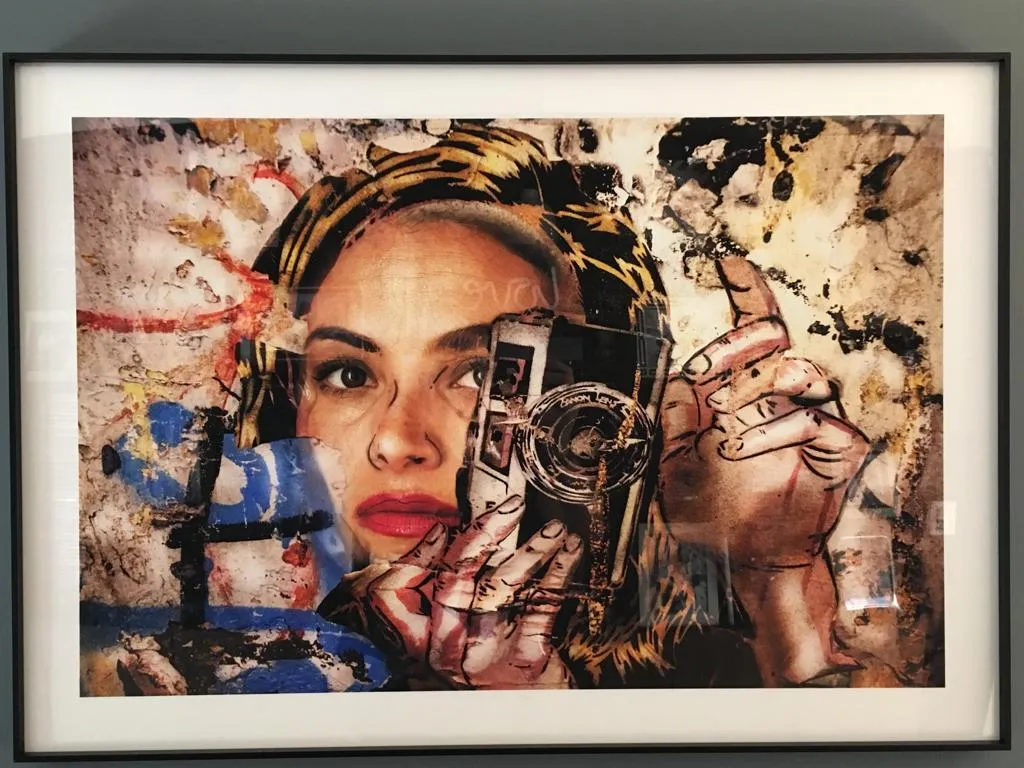 Obra de Valeria impresa y enmarcada: retrato de una mujer sosteniendo una cámara entre texturas de color, colgada en una pared