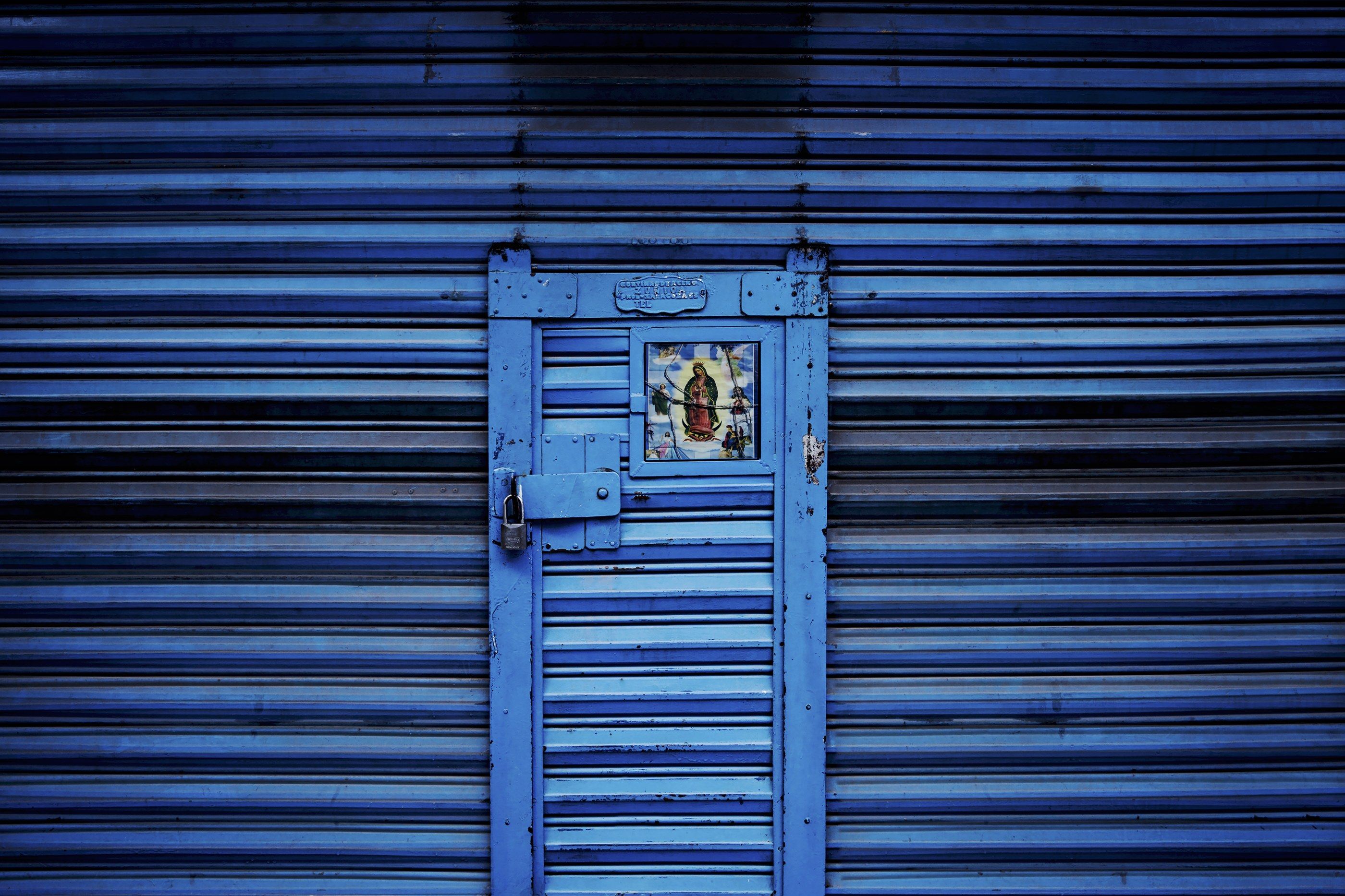 Cortina metálica azul intenso con una puerta pequeña y una estampa religiosa en el centro